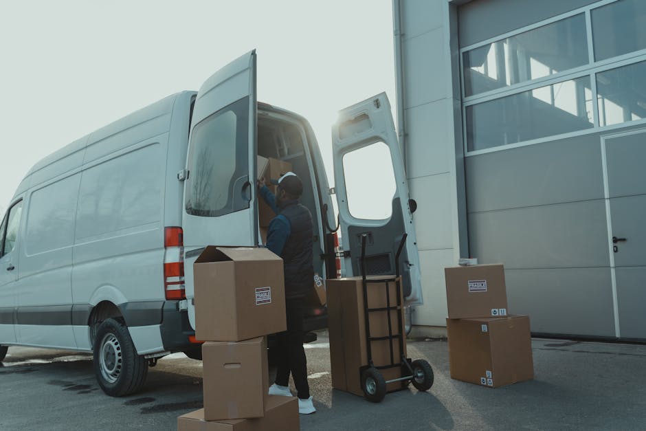 A man wearing a black cap, yellow vest, and grey trousers is bending down to lift and load a cardboard box into the open back of a white van parked on a driveway. The van is positioned next to a house