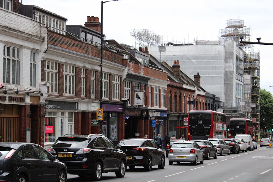 A street scene in Clapham with a row of brick terraced houses and retail shops, some of which have shopfront signage and large windows. The buildings are of varying heights and styles, with tiled roofs and brick facades. Several cars, including a black hatchback, a silver sedan, and a dark-colored SUV, are parked along the curb, with additional vehicles such as a white van and a black car waiting in traffic. Two red double-decker buses are visible, one approaching and the other stationary. Construction scaffolding covered in white sheeting is erected on the upper floors of a building at the far end of the street, indicating ongoing building work. Pedestrians are walking along the pavement, some near the shops, while others appear to be waiting or crossing the road. The scene is captured during daylight with overcast weather, lending a neutral, practical atmosphere typical of urban home relocation or moving activities, underpinning the services offered by Man with Van Clapham in house removals and furniture transport.