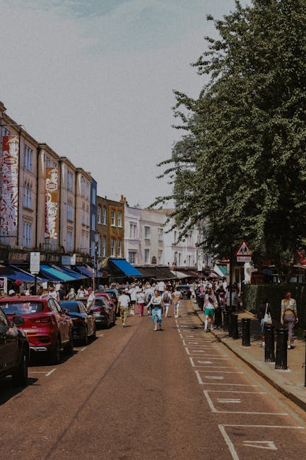 A street scene in Clapham with a row of brick terraced houses and retail shops, some of which have shopfront signage and large windows. The buildings are of varying heights and styles, with tiled roofs and brick facades. Several cars, including a black hatchback, a silver sedan, and a dark-colored SUV, are parked along the curb, with additional vehicles such as a white van and a black car waiting in traffic. Two red double-decker buses are visible, one approaching and the other stationary. Construction scaffolding covered in white sheeting is erected on the upper floors of a building at the far end of the street, indicating ongoing building work. Pedestrians are walking along the pavement, some near the shops, while others appear to be waiting or crossing the road. The scene is captured during daylight with overcast weather, lending a neutral, practical atmosphere typical of urban home relocation or moving activities, underpinning the services offered by Man with Van Clapham in house removals and furniture transport.