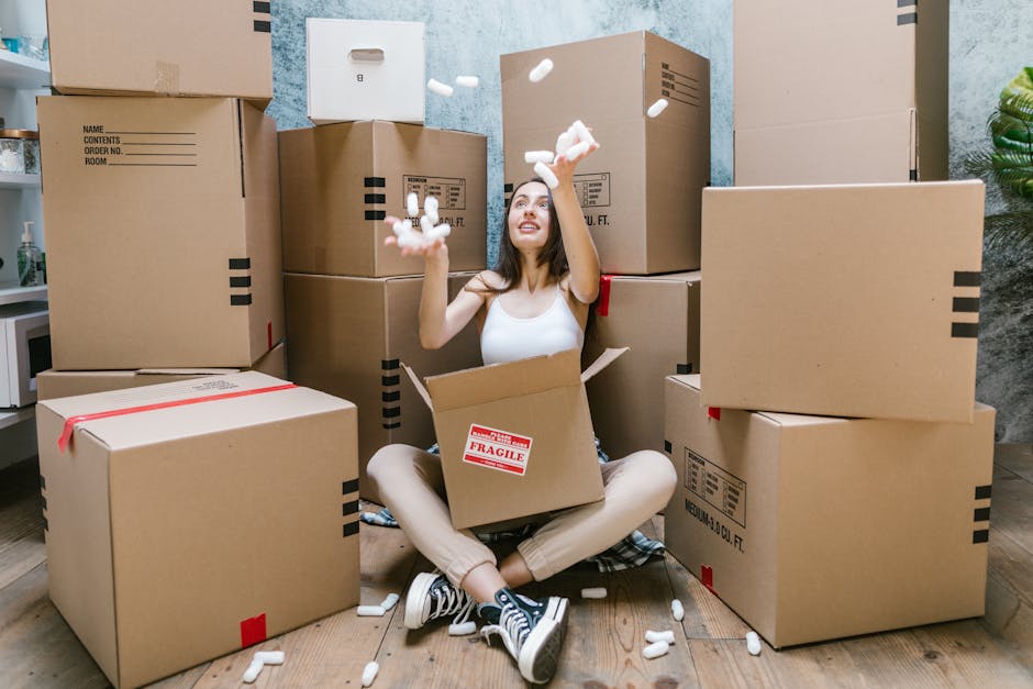 A young woman with long brown hair is sitting cross-legged on a wooden floor inside a room during a home relocation process. She is surrounded by numerous large cardboard moving boxes, some stacked on top of each other and others placed directly on the floor. The woman is wearing a white sleeveless top, beige pants, and black-and-white sneakers, and has a cheerful expression as she tosses white packing foam sheets into the air. An open cardboard box with a red 'Fragile' sticker rests on her lap, while more foam packing materials are scattered around her. The room’s background features textured blue-gray walls, a small potted plant to the right, and a glimpse of a kitchen countertop with a plastic bottle on the left. This scene appears to depict the packing and loading process associated with furniture transport and home removals, as handled by [COMPANY_NAME], in preparation for transportation as part of a professional house moving service.