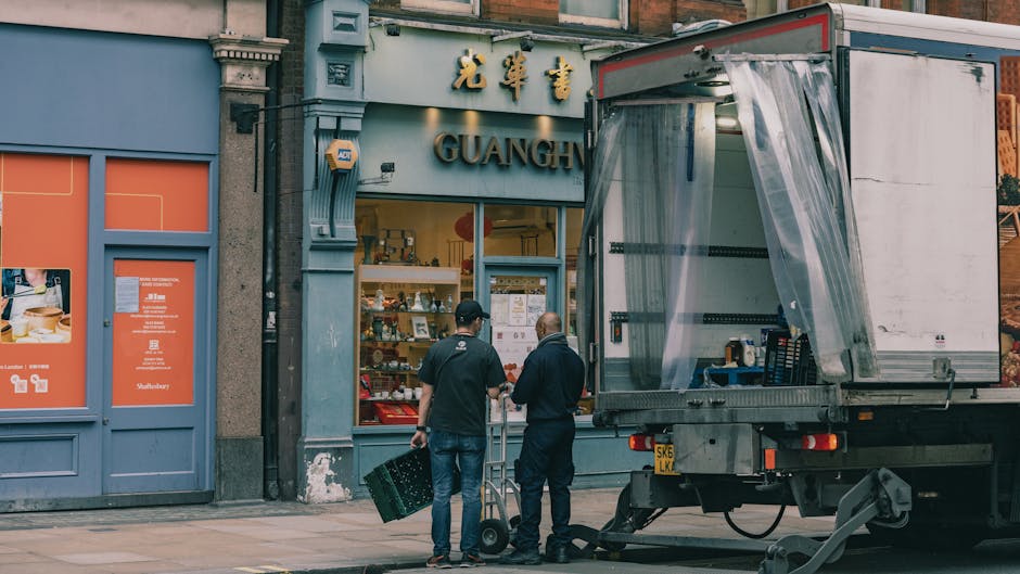 Two men are preparing to load or unload a large moving van parked on the pavement in front of a shop with a blue and orange exterior. One man, dressed in dark clothing, is carrying a green plastic crate, while the other, also in dark clothing, is standing nearby with a hand on a hand truck or dolly. The open side of the van reveals a plastic sheet covering the interior, with some shelves, boxes, and packing materials visible inside. The shop behind them has large windows displaying various items and a sign with Asian characters and the word 'GUANG' visible above the entrance. The scene captures the process of home relocation or furniture transport as part of a house removal service, with materials like cardboard boxes and packing equipment present, emphasizing the loading or packing stage of a move. Man with Van Clapham provides this type of furniture transport and packing service, supporting smooth relocation logistics.