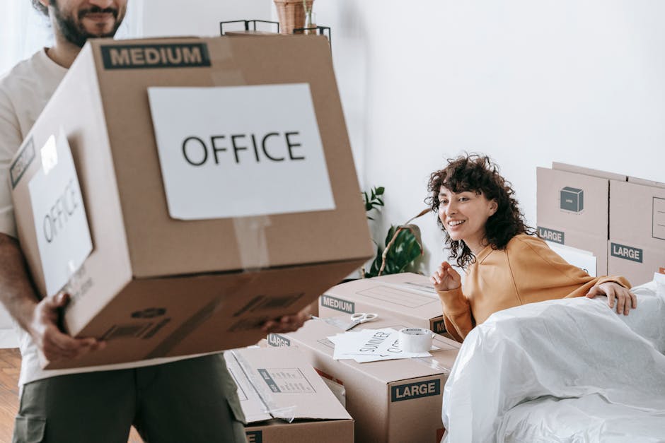 A young woman with long brown hair is sitting cross-legged on a wooden floor inside a room during a home relocation process. She is surrounded by numerous large cardboard moving boxes, some stacked on top of each other and others placed directly on the floor. The woman is wearing a white sleeveless top, beige pants, and black-and-white sneakers, and has a cheerful expression as she tosses white packing foam sheets into the air. An open cardboard box with a red 'Fragile' sticker rests on her lap, while more foam packing materials are scattered around her. The room’s background features textured blue-gray walls, a small potted plant to the right, and a glimpse of a kitchen countertop with a plastic bottle on the left. This scene appears to depict the packing and loading process associated with furniture transport and home removals, as handled by [COMPANY_NAME], in preparation for transportation as part of a professional house moving service.